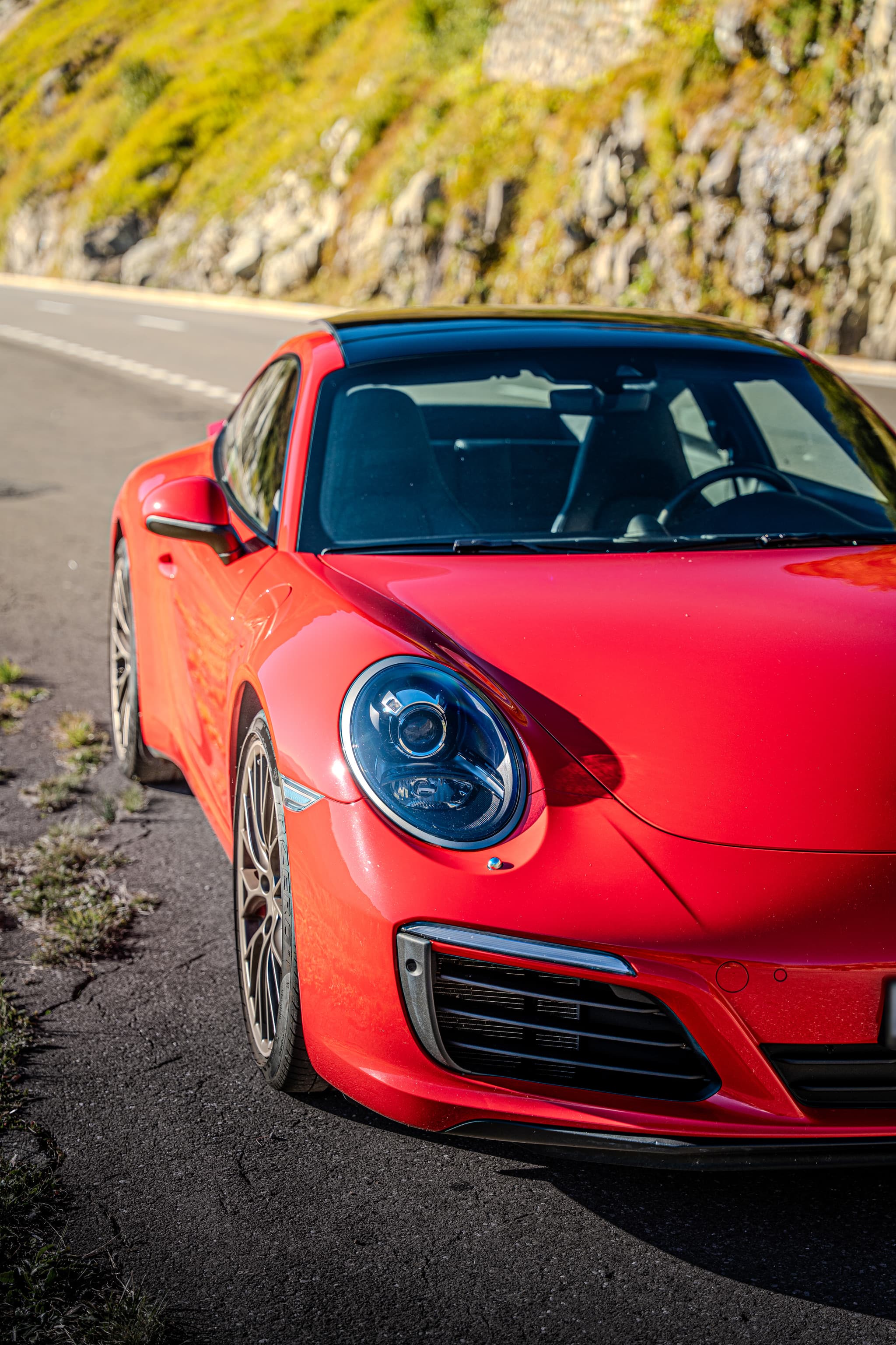Red Porsche 911 Carrera S Coupé, headlight and nose in close-up at the roadside, a green slope behind