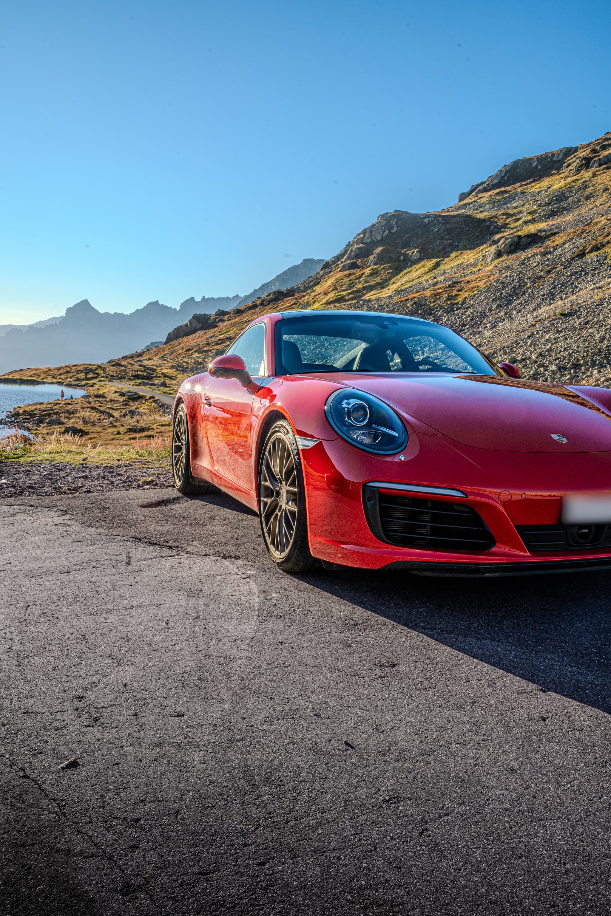 Red Porsche 911 Carrera S Coupé, close front three-quarter view, a mountain lake on the left and a sunlit scree slope behind