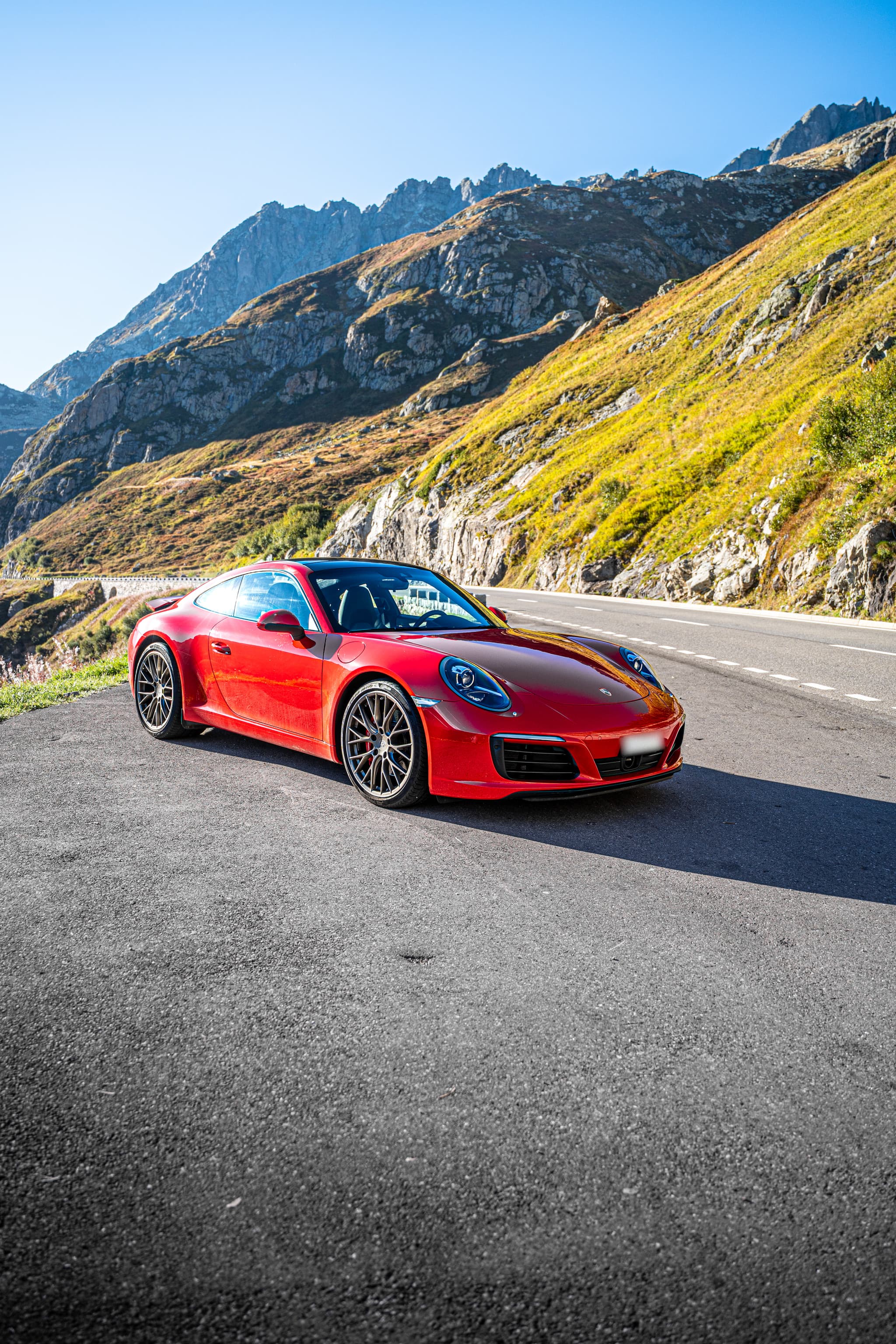 Red Porsche 911 Carrera S Coupé, front three-quarter view on a pass road between rocks and sunlit mountainsides