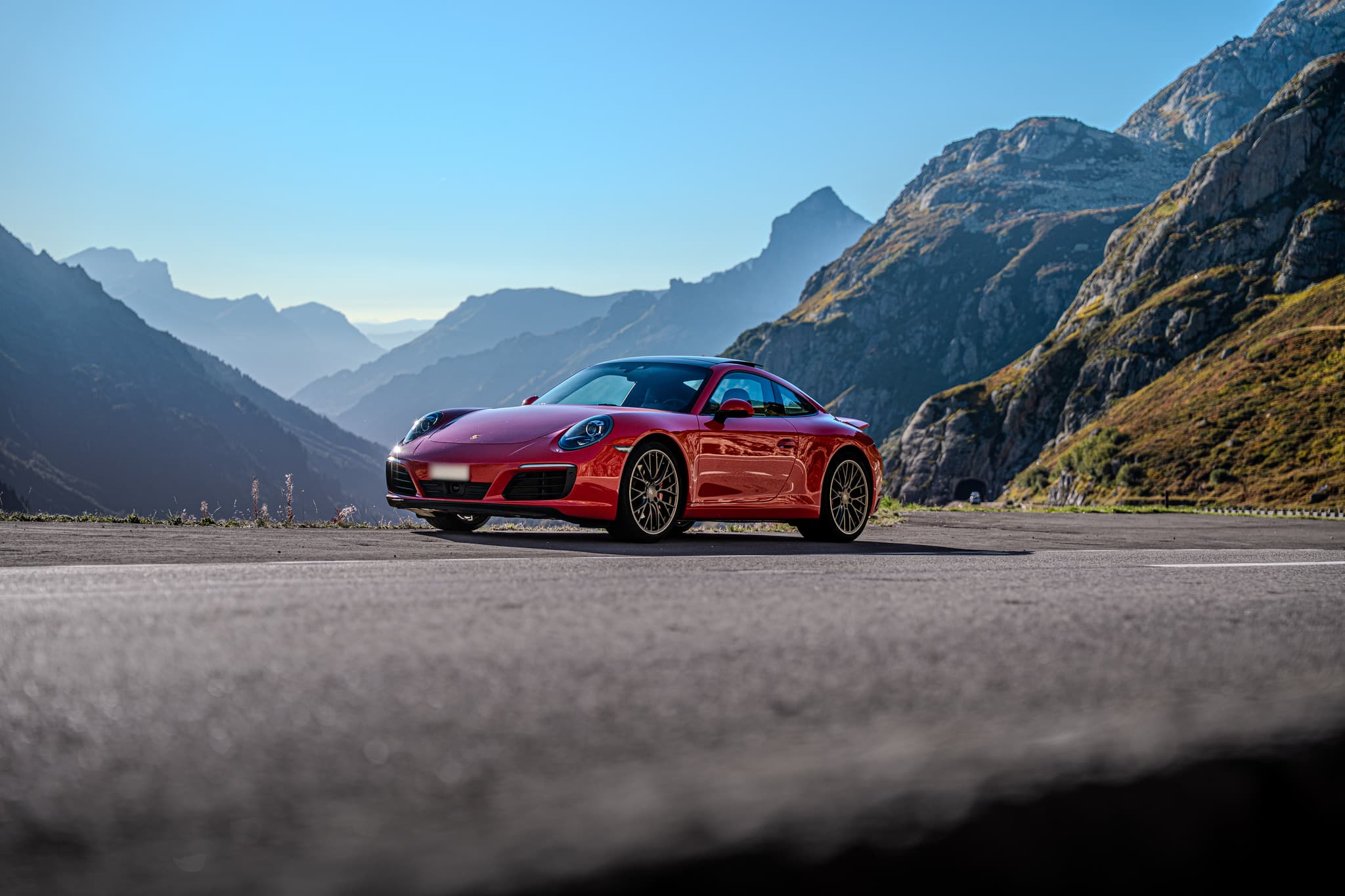 Red Porsche 911 Carrera S Coupé, front three-quarter view on a pass road, layered mountain ridges against the light behind
